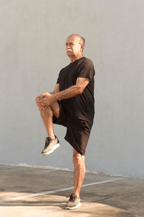 Eine Person in schwarzem T-Shirt und schwarzen Shorts steht auf einem Bein und zieht das angewinkelte Knie mit beiden Händen zur Brust, eine Dehn- oder Kräftigungsübung ausführend.