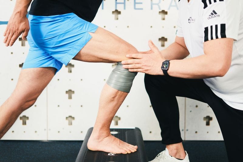 Ein Physiotherapeut in weißem Adidas-Shirt führt an einer Person in blauen Shorts Medical Flossing am Knie durch, wobei das Knie mit einem grauen Flossband umwickelt ist und die Person auf einem Therapiestep steht.