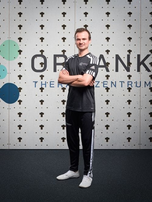 Kursleiter Jakob steht mit verschränkten Armen vor einer Holzlochplatten-Wand mit dem Schriftzug „Orbanke Therapiezentrum" und trägt schwarze Adidas-Sportkleidung mit weißen Streifen.