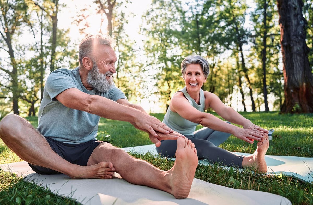 Flexible Übungen für den Körper. Sportliche Männer und Frauen mit grauem Haar dehnen sich beim Training im Freien auf Yogamatten mit den Händen an einem Bein. Glückliches Ehepaar mit nackten Füßen, das sich gemeinsam im Park aufwärmt.