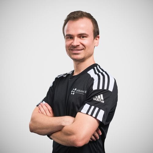 Porträtfoto von Kursleiter Jakob vor hellem Hintergrund, mit verschränkten Armen und freundlichem Lächeln, gekleidet in ein schwarzes Adidas-Sportshirt mit dem Logo der Physiotherapie Orbanke.